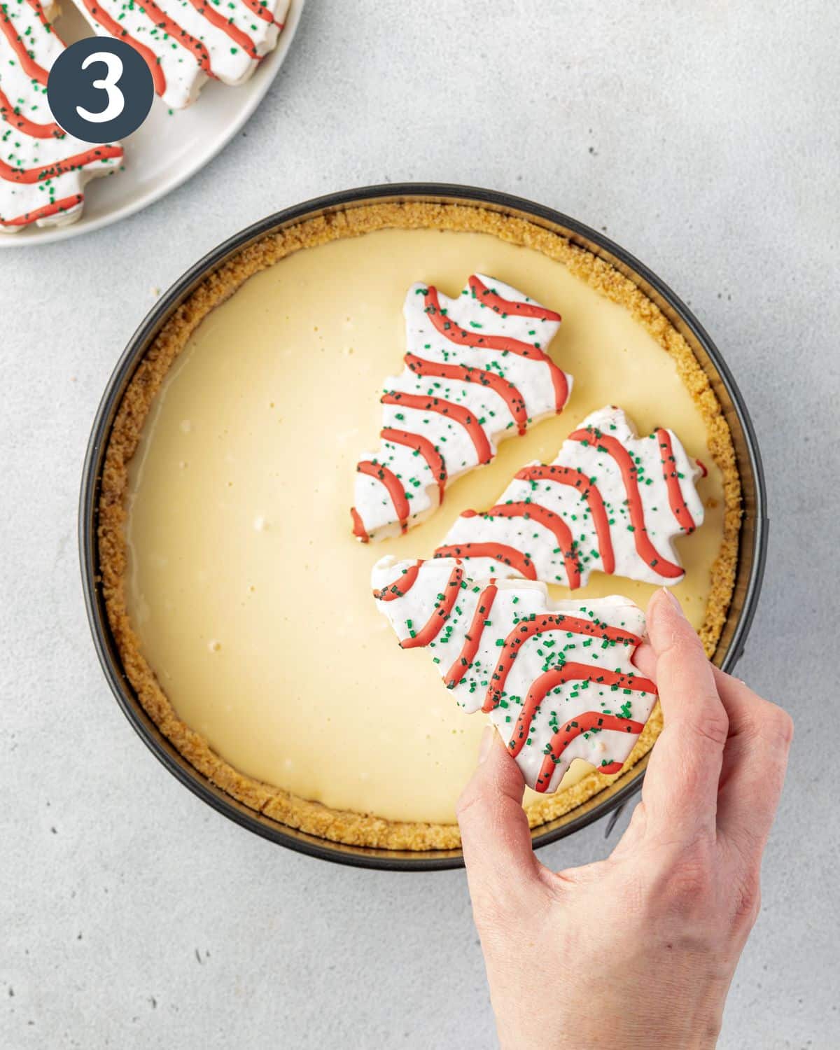 2 Little Debbie Christmas tree cakes in uncooked cheesecake batter with tops towards the center, and a hand placing a third tree in.
