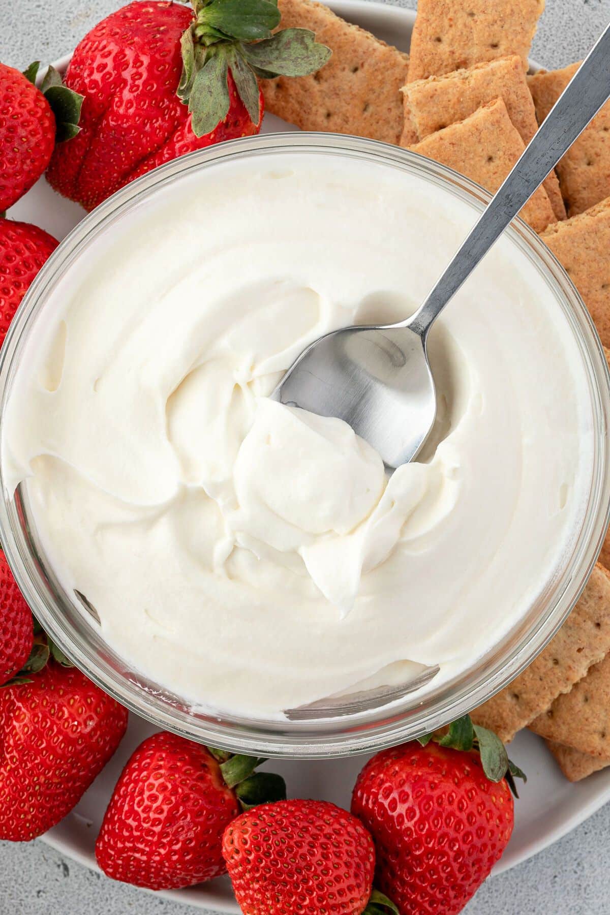 Marshmallow whipped cream in a bowl with a silver spoon on a platter of strawberries and graham crackers.