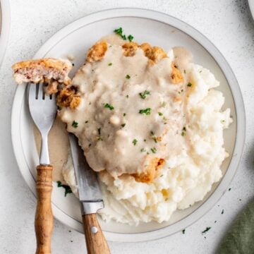 A round plate with mashed potatoes and chicken fried steak topped with gravy, and a fork and knife with a bite on the side.