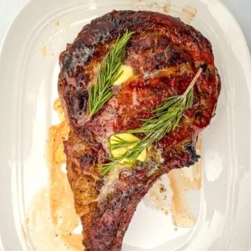 Bone-in ribeye topped with pats of butter and sprigs of rosemary on a white oval platter.