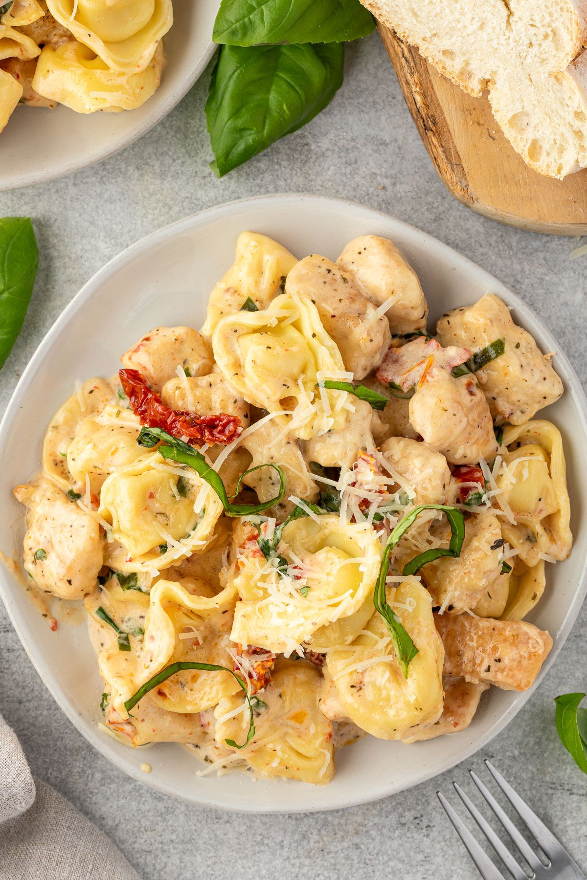 A serving of cheese tortellini and chicken in a marry me sauce on a plate, with bread in one corner and another serving on a plate in another.