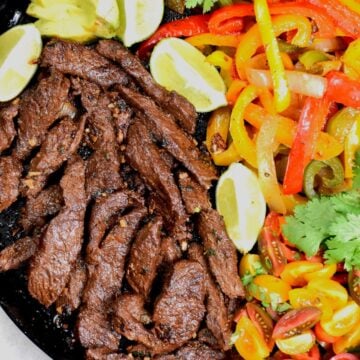 A cast iron skillet with strips of fajita meat, multi-colored peppers, lime wedges and cilantro.