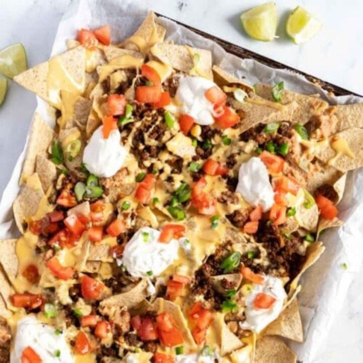 A parchment-lined sheet pan filled with beef nachos and topped with green onions, diced tomatoes, and scoops of sour cream.