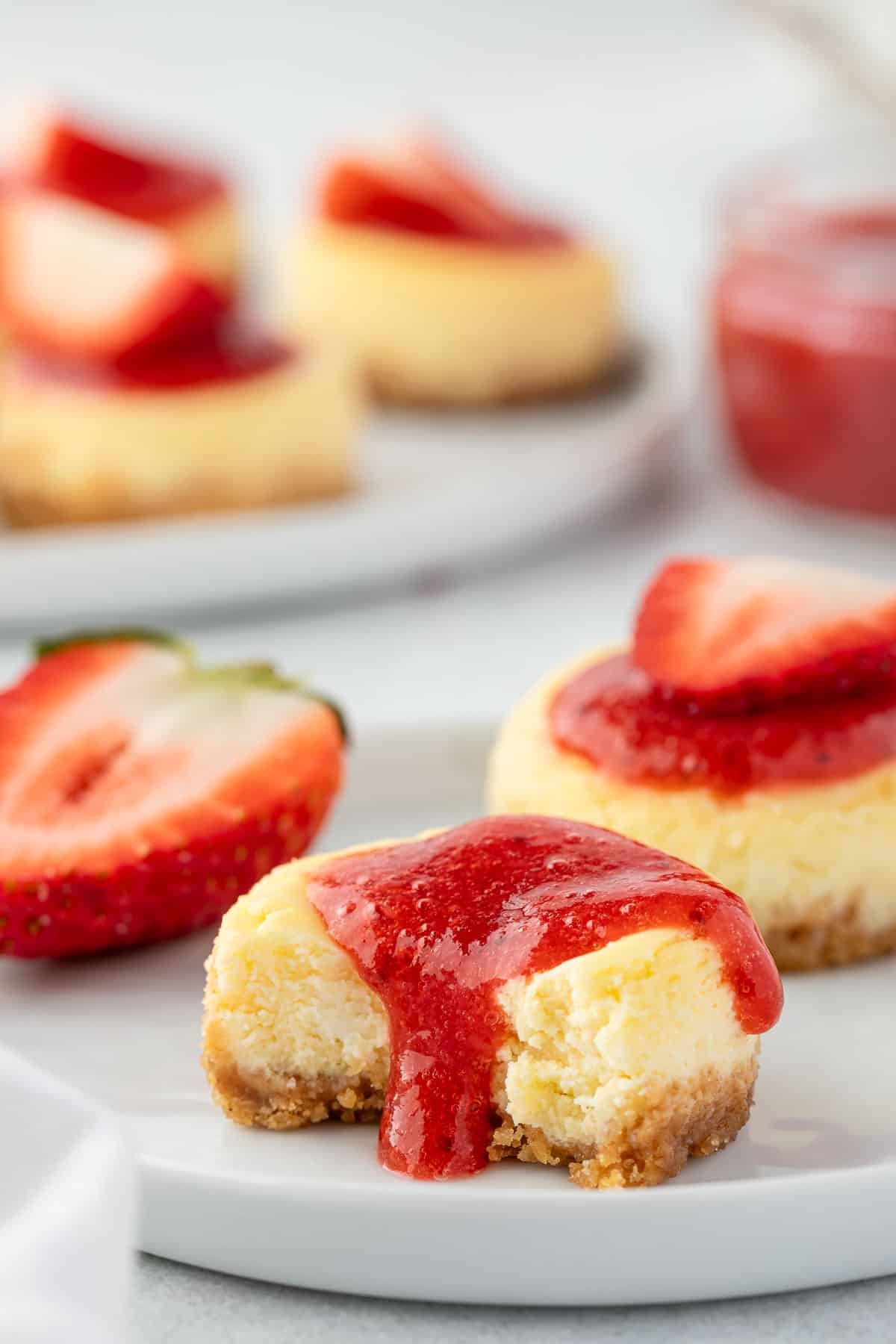 Miniature strawberry cheesecake with a bite missing and strawberry sauce dripping down the sides.