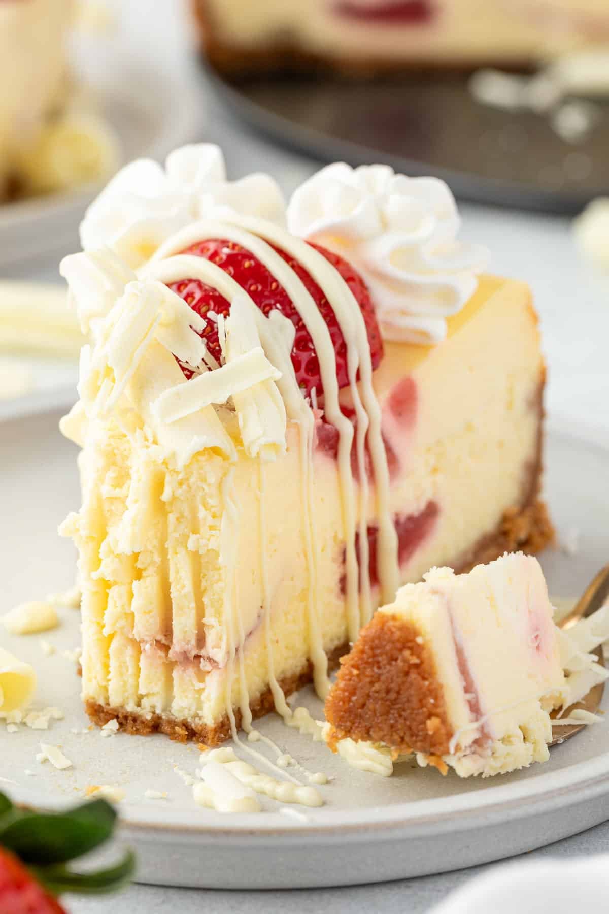 A slice of strawberry white chocolate cheesecake with a bite missing, topped with fresh strawberries and a drizzle of chocolate sauce.