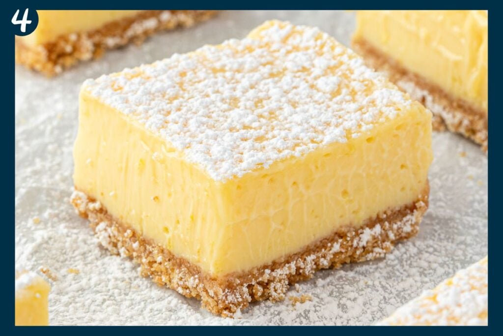 A lemon bar with a crumbly crust, creamy filling, and powdered sugar on top, shown up close.