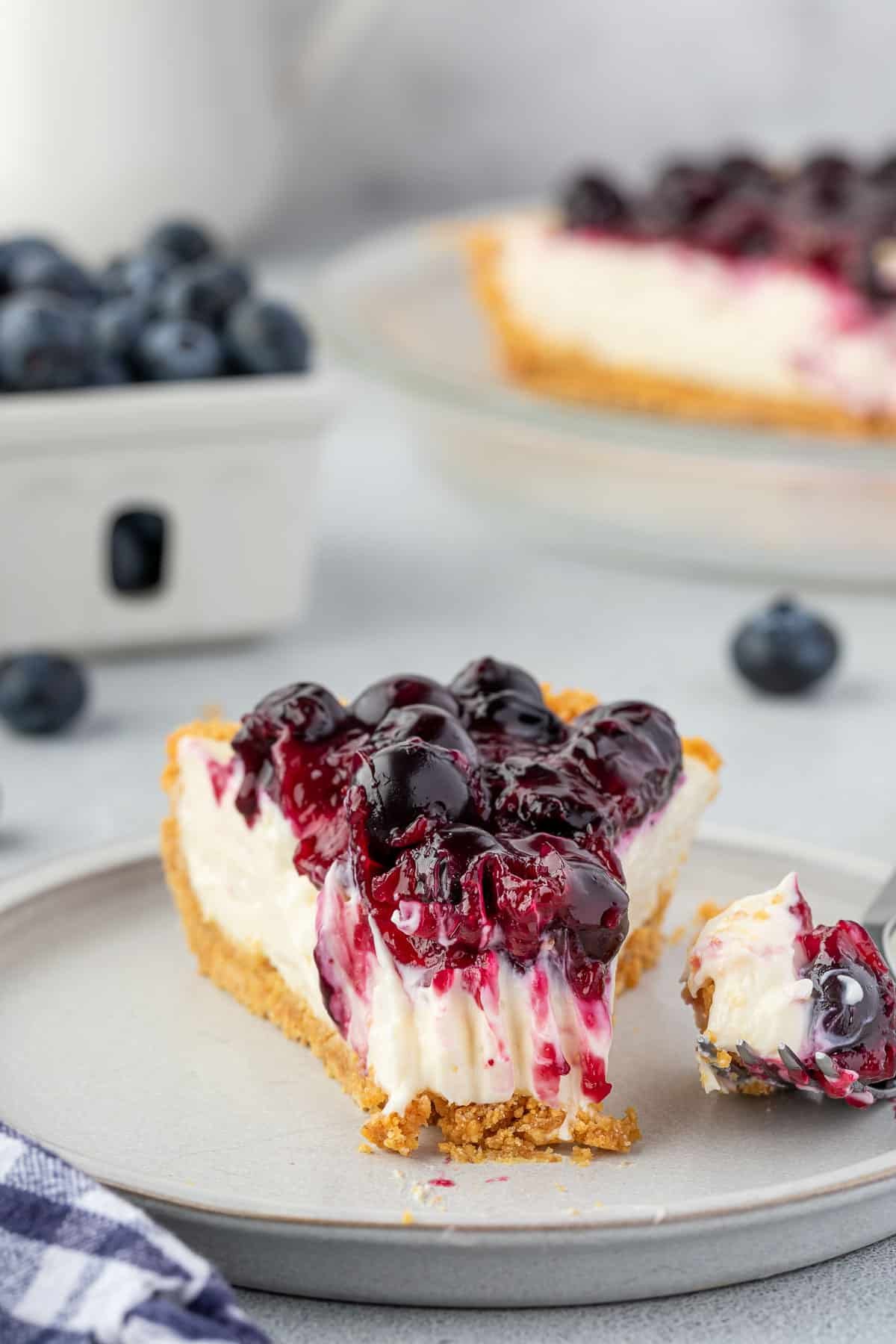 Slice of blueberry cream cheese pie on a plate with a bite on a fork, and a ceramic basket filled with blueberries.