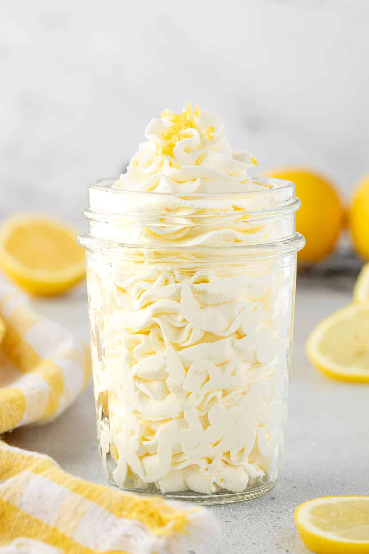 Lemon whipped cream piped in a glass jar with sliced lemons and a yellow checkered towel around it.