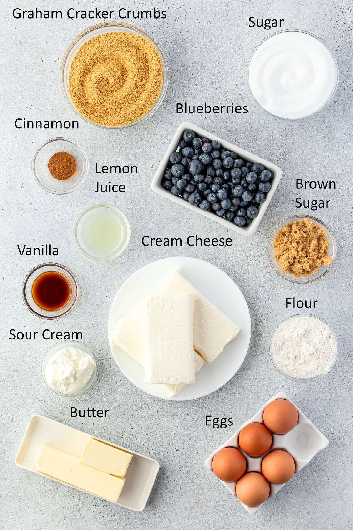 Recipe ingredients neatly arranged on a gray surface and labeled, items include graham cracker crumbs, sugar, blueberries, brown sugar, flour, eggs, cream cheese, butter, whipped cream, vanilla, lemon juice, sour cream, and cinnamon.