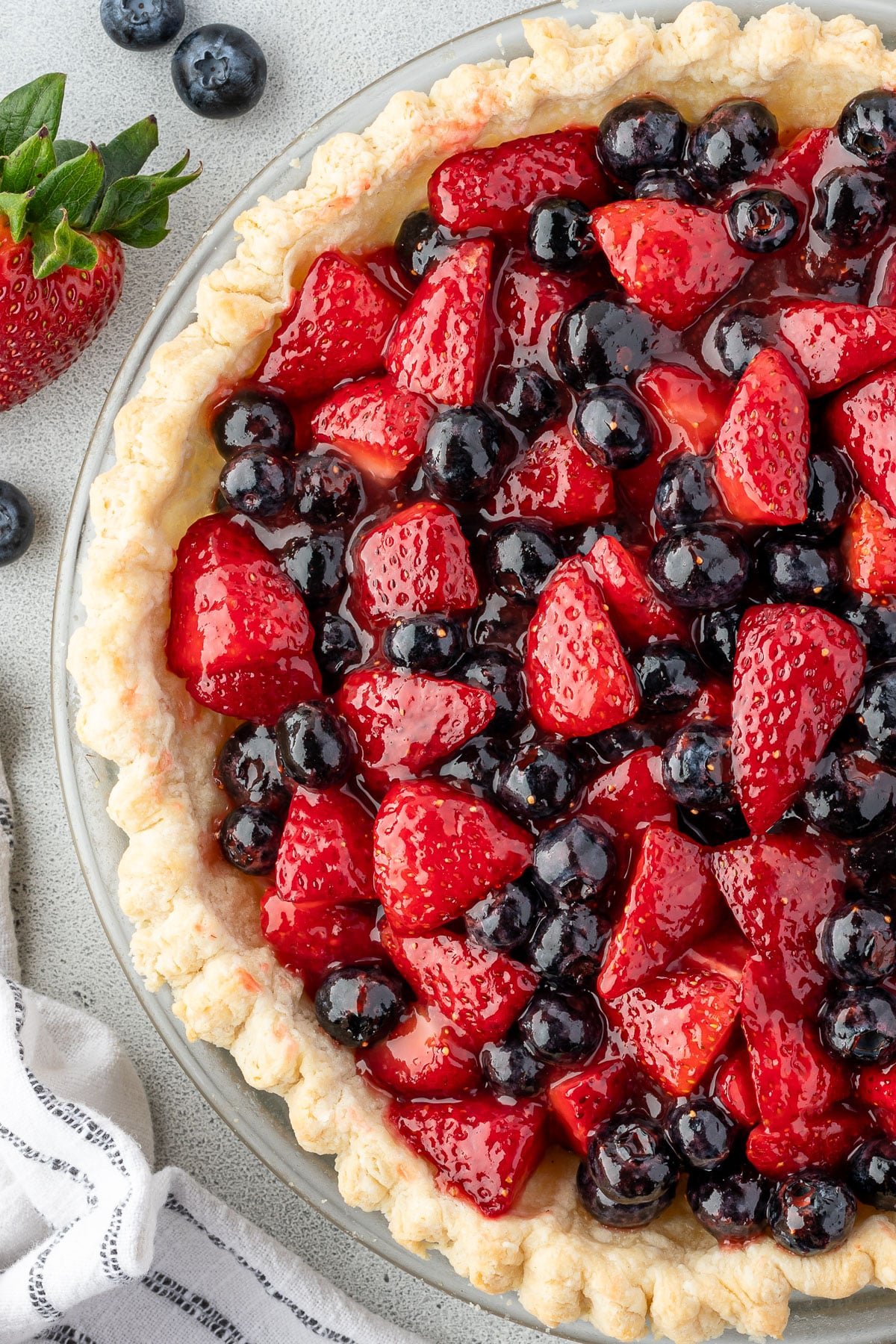 Close-up of a fresh strawberry and blueberry pie with glossy fruit in a flaky crust, with a linen beside it.