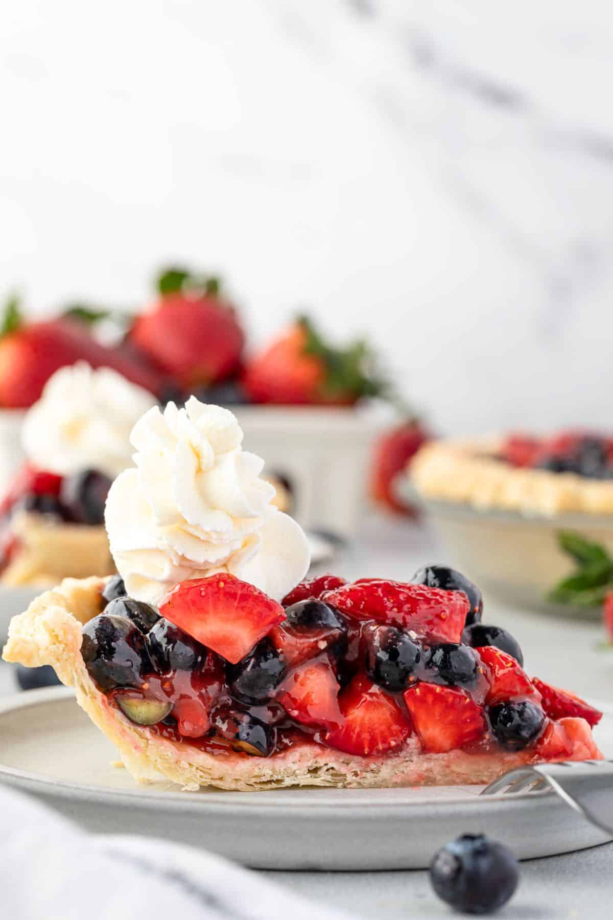 A slice of blueberry strawberry pie topped with whipped cream on a plate, with whole strawberries and blueberries in the background.