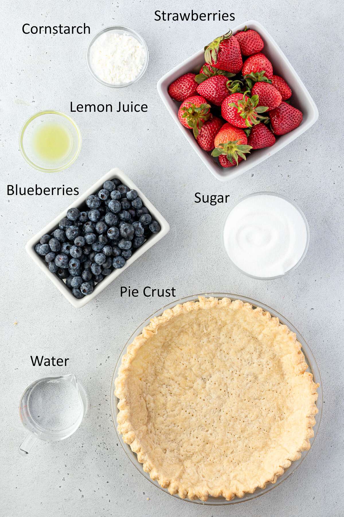 Top-down view of ingredients for making a pie, including strawberries, blueberries, cornstarch, lemon juice, sugar, water, and a pre-made pie crust, all neatly labeled on a white surface.