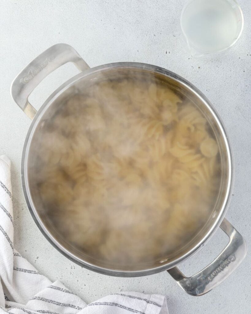 A stainless steel pot on a grey countertop filled with boiling water and fusilli pasta with steam above the pan.
