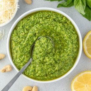 A white bowl filled with basil cashew pesto with a spoon resting it in and some ingredients scattered around the bowl.