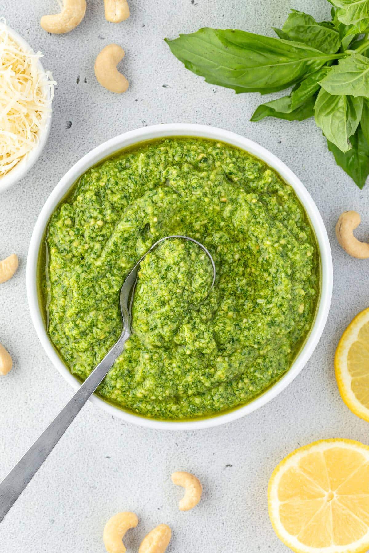 A bowl of vibrant green basil cashew pesto with a spoon in it surrounded by lemon slices, cashews, fresh basil leaves, and a small bowl of shredded cheese.