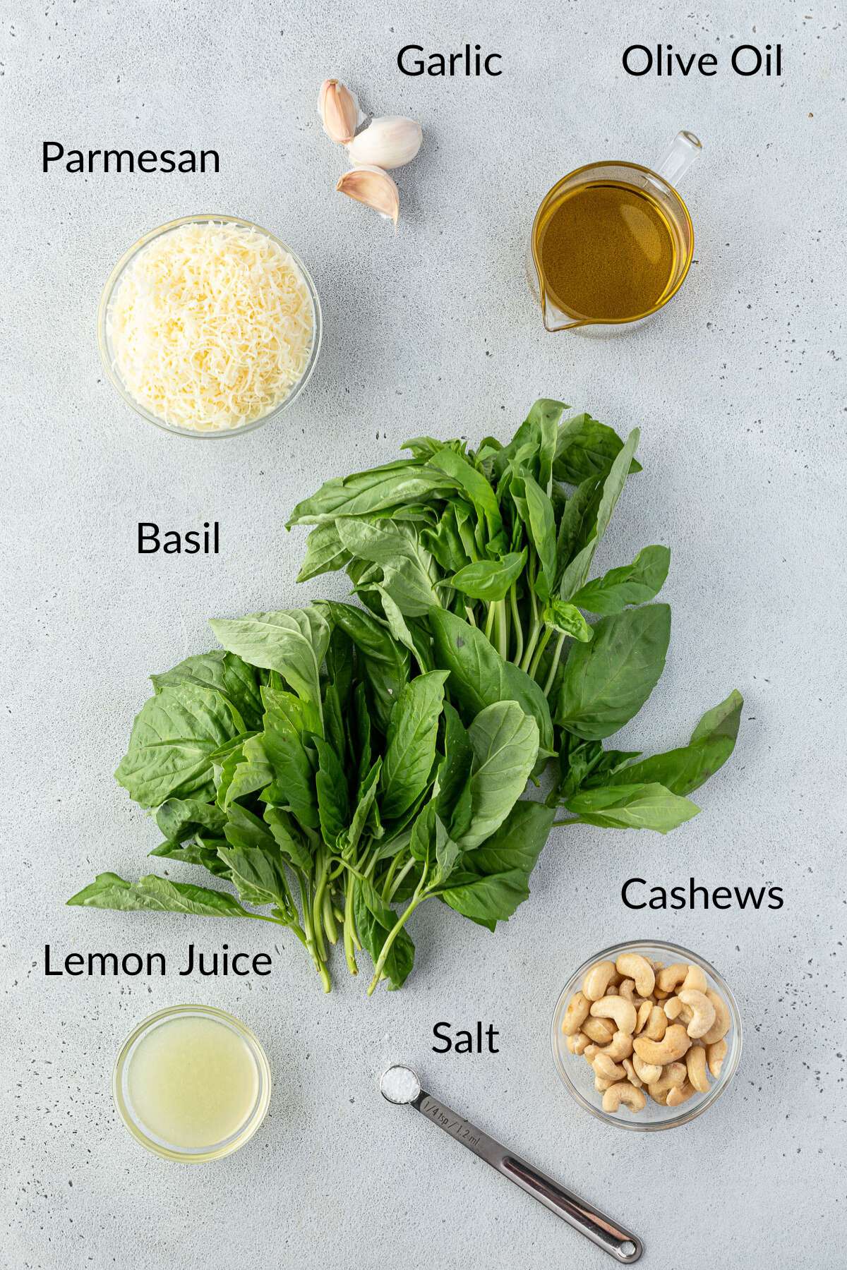 A flat lay of ingredients for making pesto displayed on a light surface with the items in labeled bowls.