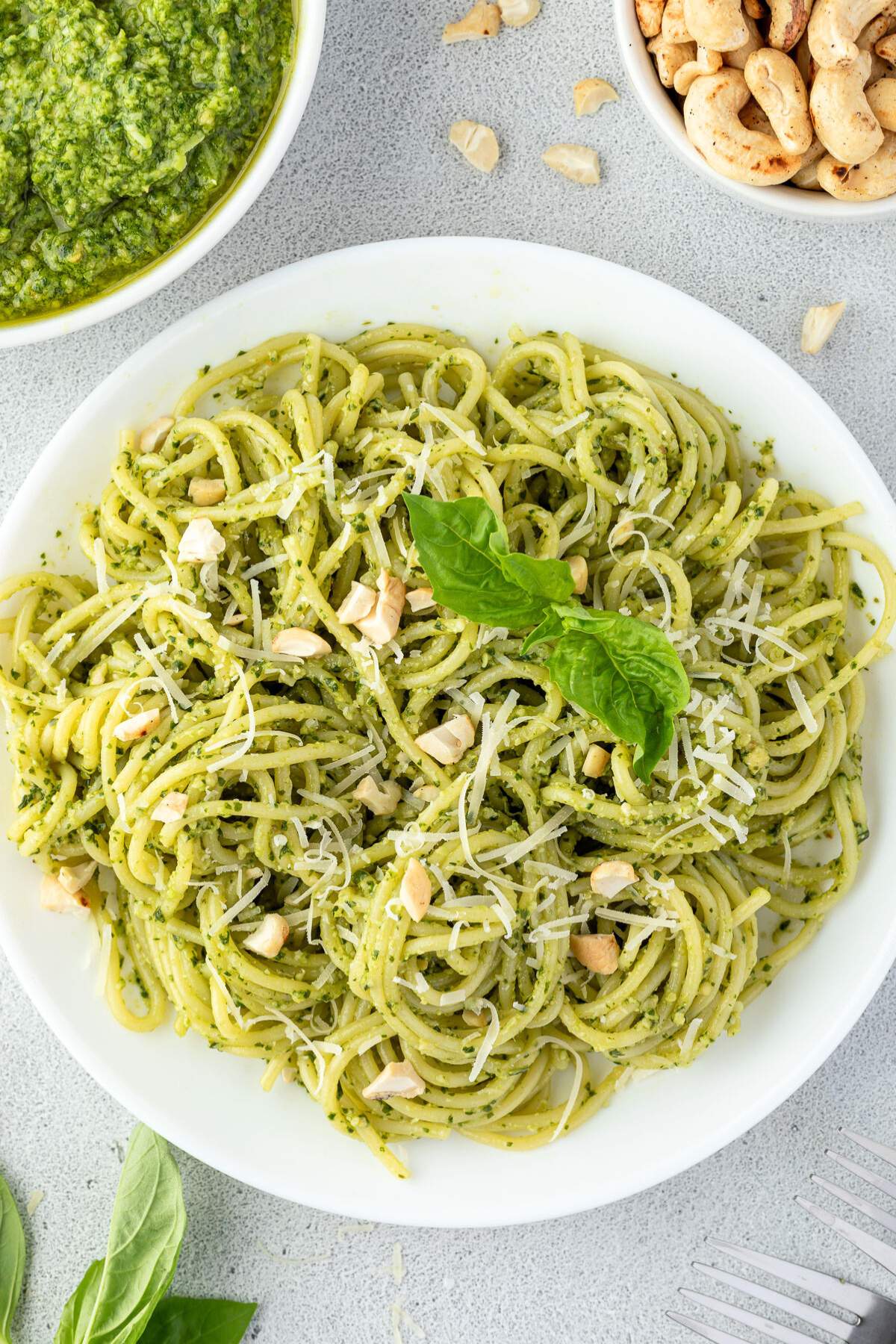 A plate of spaghetti pesto garnished with grated cheese, chopped nuts, and fresh basil leaves.