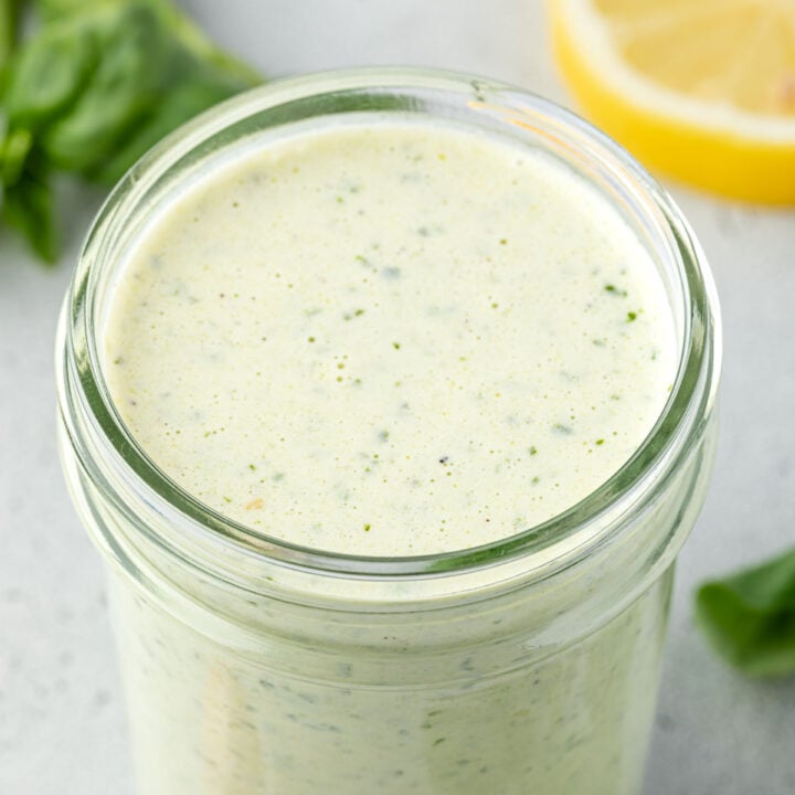 A clear glass jar filled with creamy green salad dressing sits on a light grey surface.