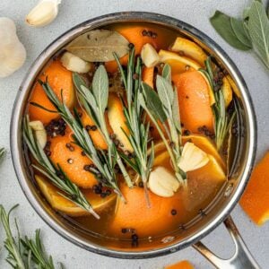 A pot, seen from above, is filled with orange slices, garlic cloves, rosemary, sage, black pepper, and bay leaves in liquid.