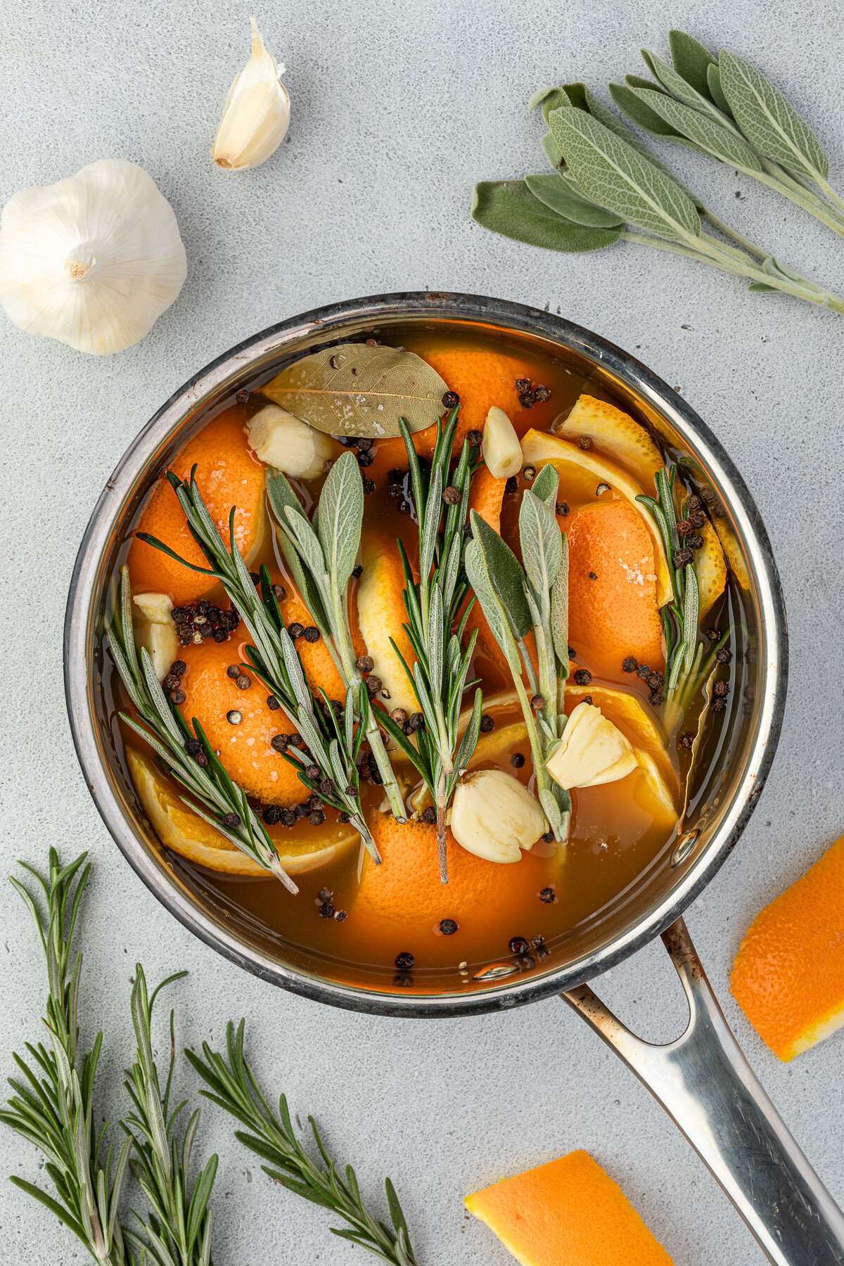 A pot with orange slices, rosemary, sage, garlic cloves, peppercorns, and bay leaves simmering in apple cider.