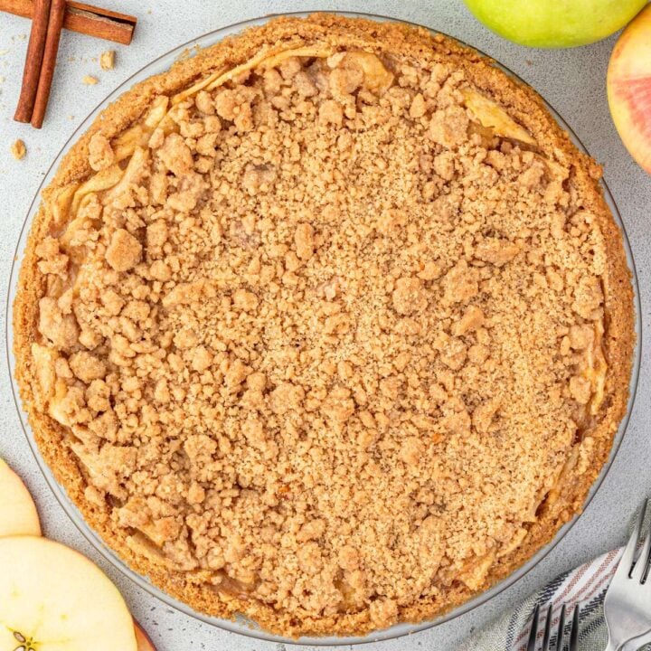 A crumb-topped apple pie on a gray surface framed with apples, cinnamon sticks, and crumbs.