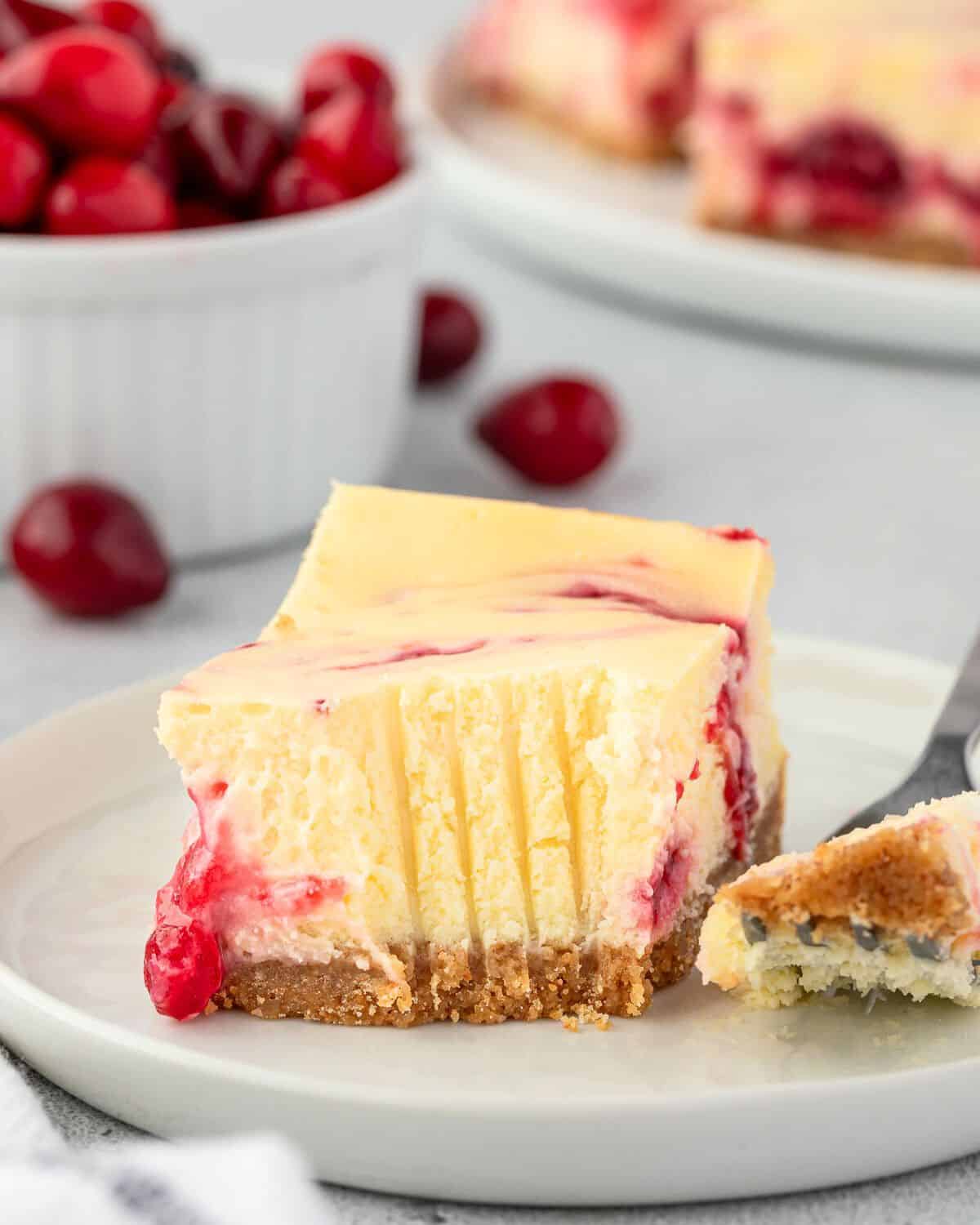 A plate with a square of cranberry cheesecake with a bite out of it, and a bowl of cranberries in the background.