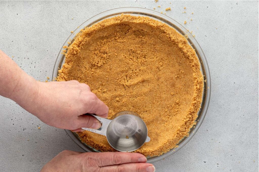 Hands pressing a no-bake graham cracker crust into a glass pie dish using a measuring cup on a gray surface.