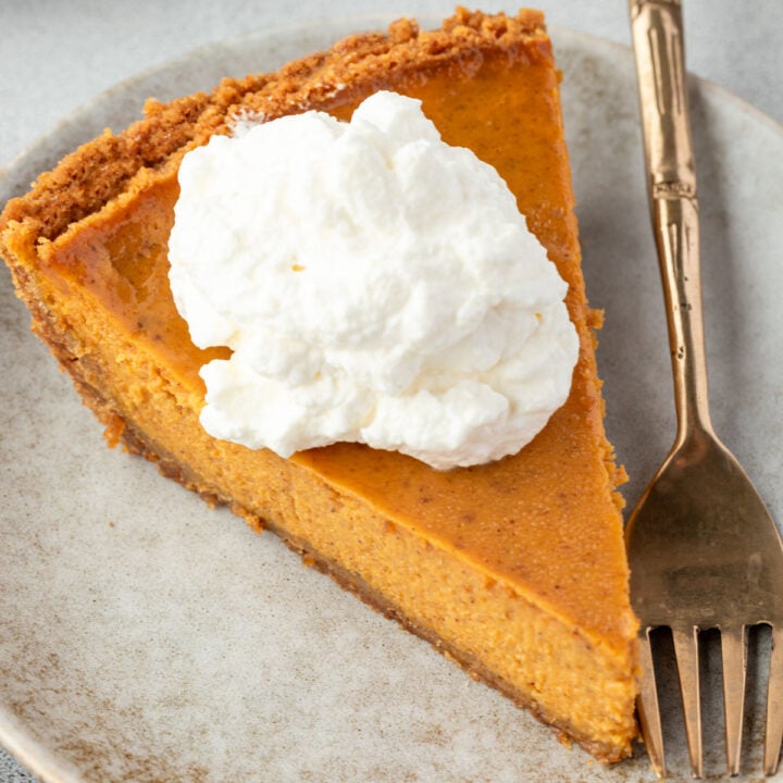 A slice of pumpkin pie with a dollop of whipped cream rests on a graham cracker crust, served on a plate with a fork.