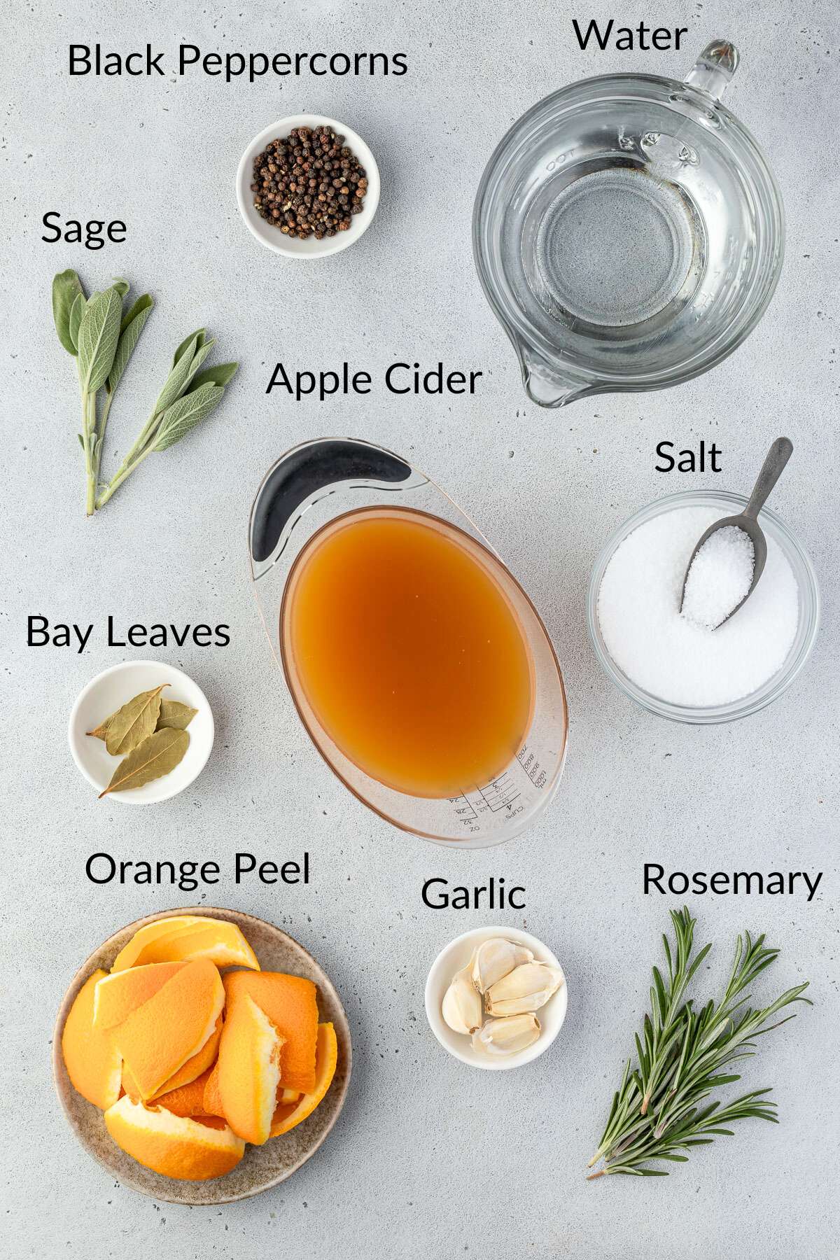 Ingredients for a turkey brine include water, apple cider, salt, black peppercorns, sage, bay leaves, orange peel, garlic, and rosemary, all arranged on a gray surface.