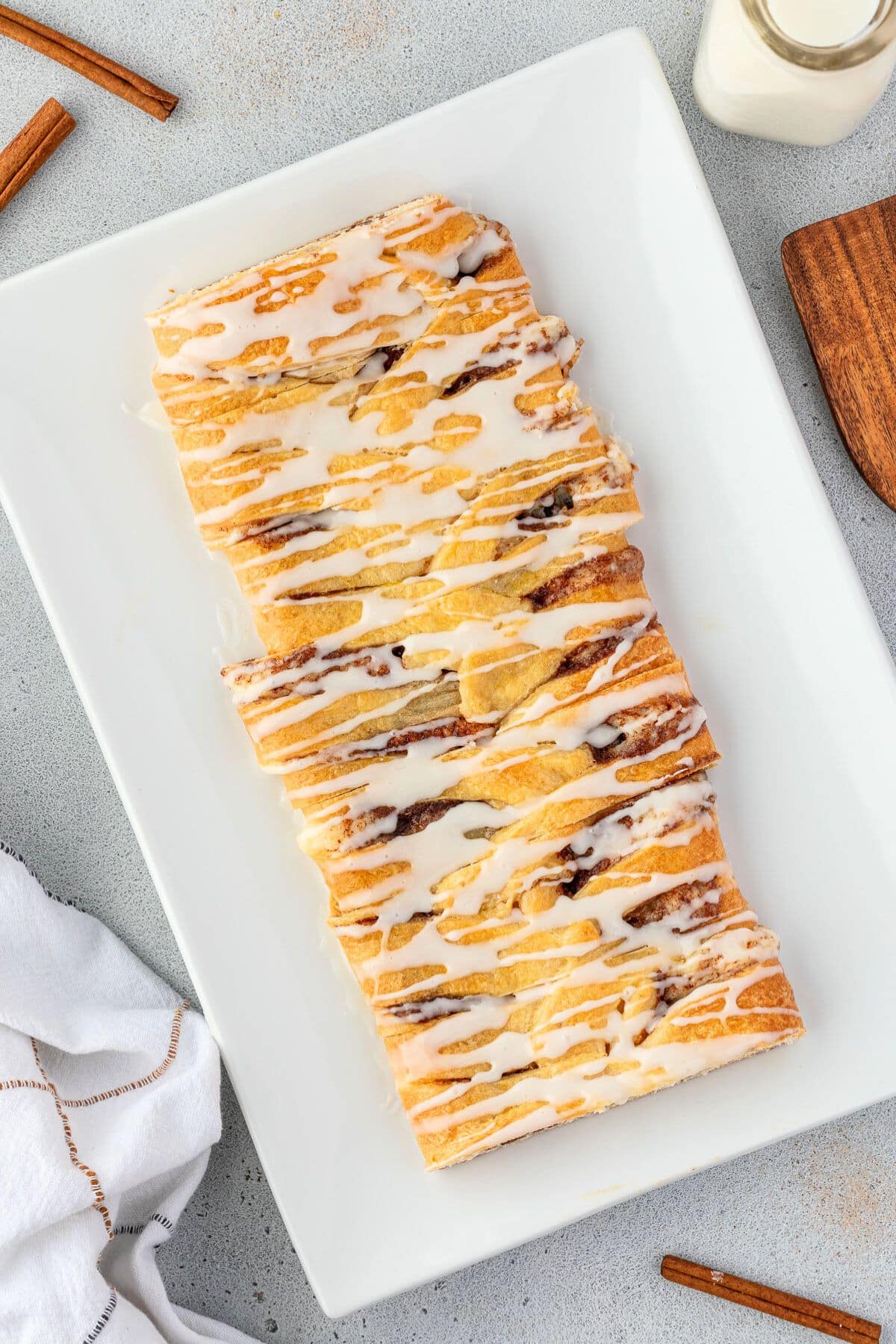 A rectangular Cinnamon Danish, drizzled with white icing, on a white rectangular plate and accompanied by a bottle of milk, a wooden utensil, and cinnamon sticks.