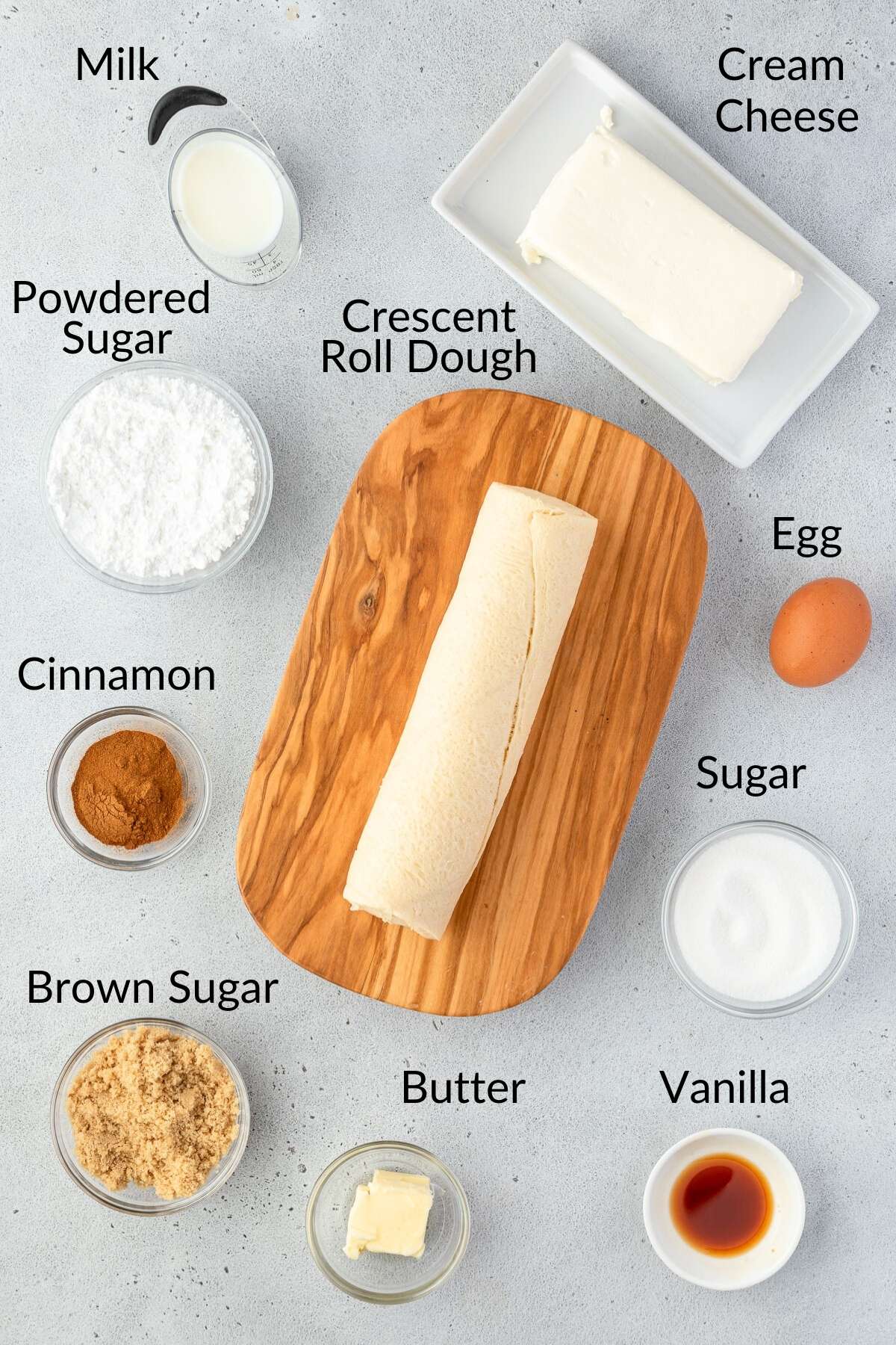 Ingredients for a simple Danish Braid are laid out on a surface, featuring cream cheese, crescent roll dough, egg, sugar, vanilla, butter, brown sugar, cinnamon, powdered sugar, and milk.