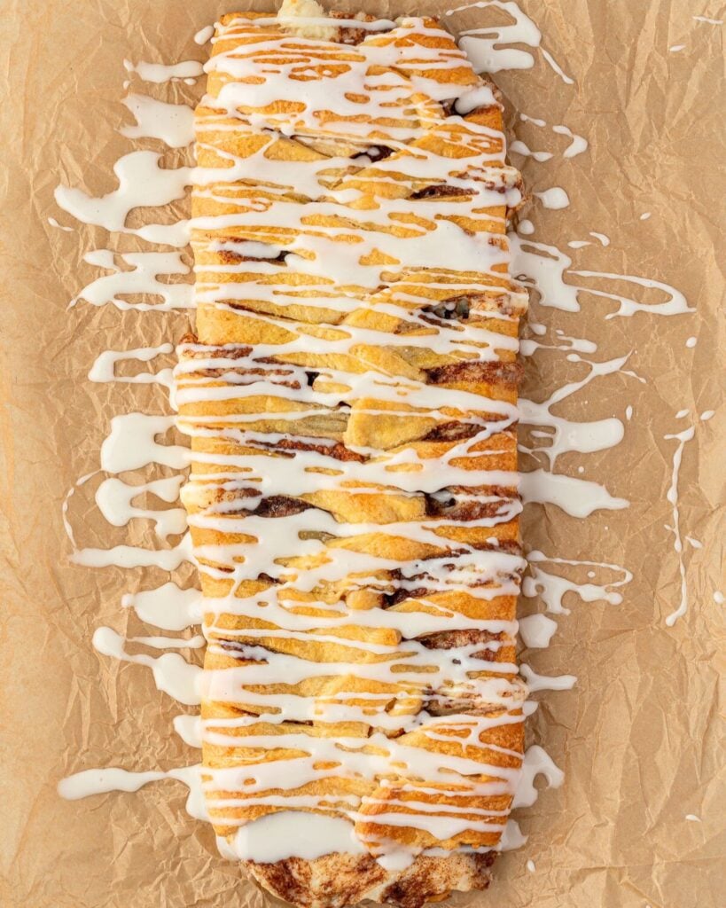 A cinnamon danish braid on crinkled parchment with glaze drizzled over it and dripping onto the paper.