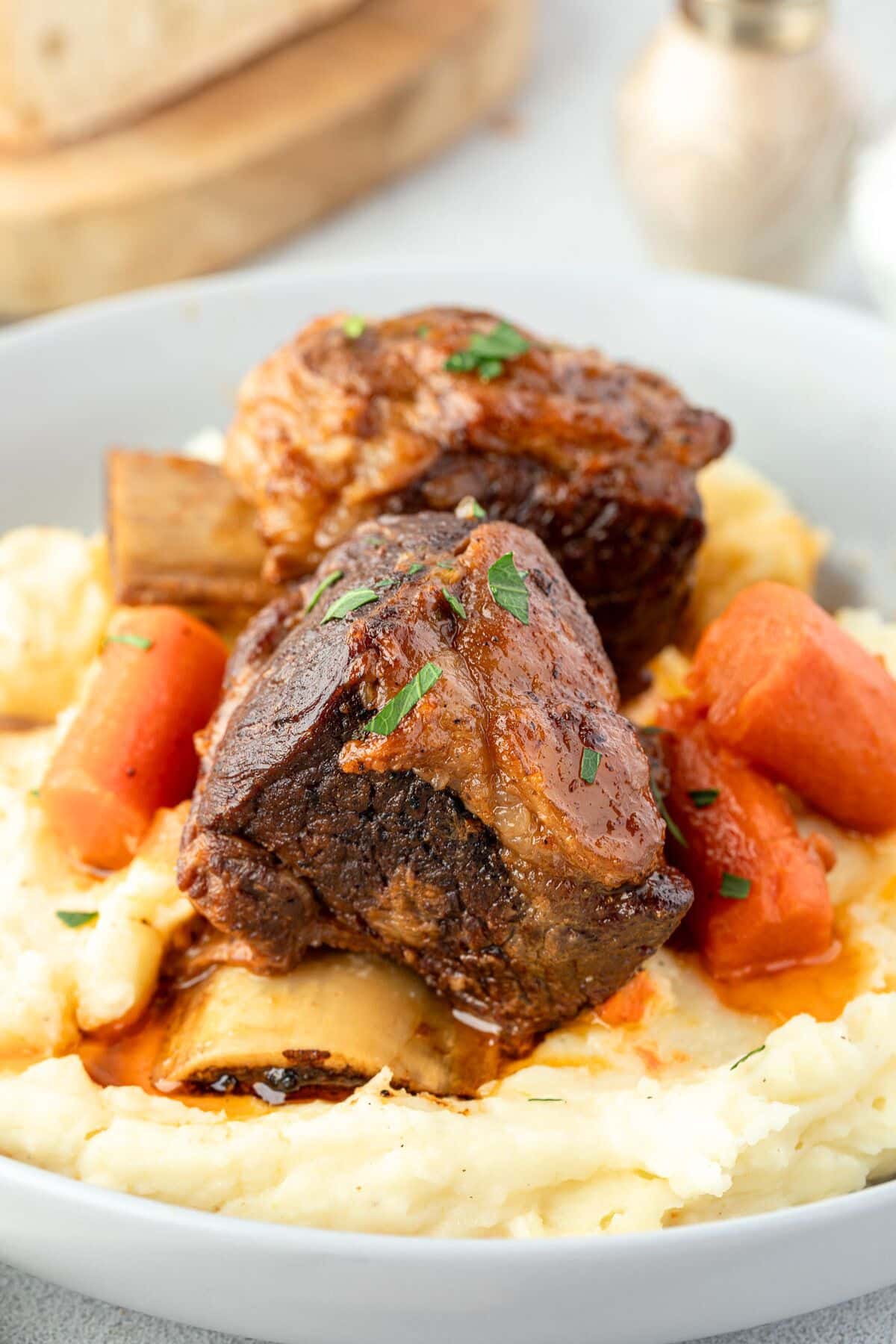 Beer-braised short ribs nestled on a bed of mashed potatoes, delicately garnished with herbs and served alongside tender carrot pieces.