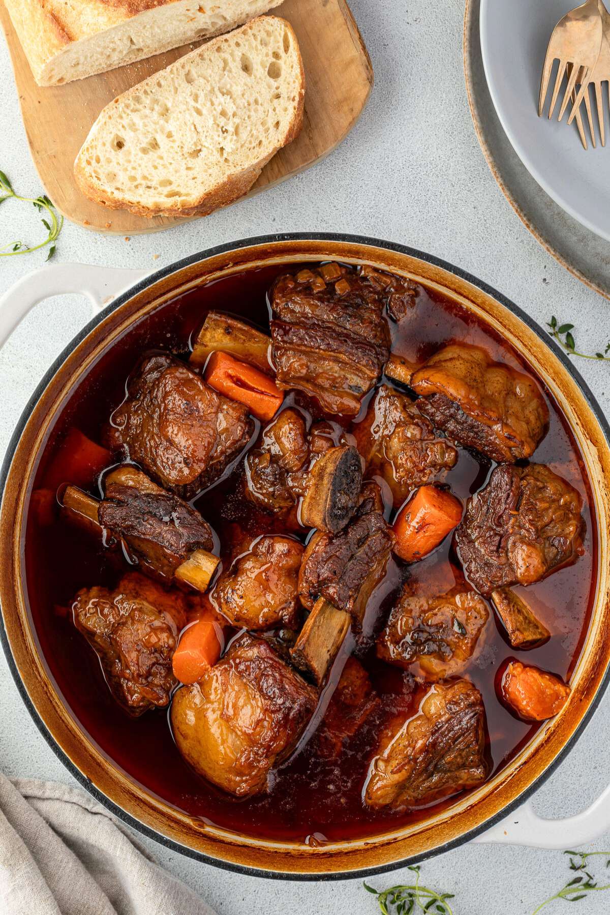 A pot of beer-braised short ribs with carrots, nestled in aDutch oven, accompanied by sliced bread on a wooden board.