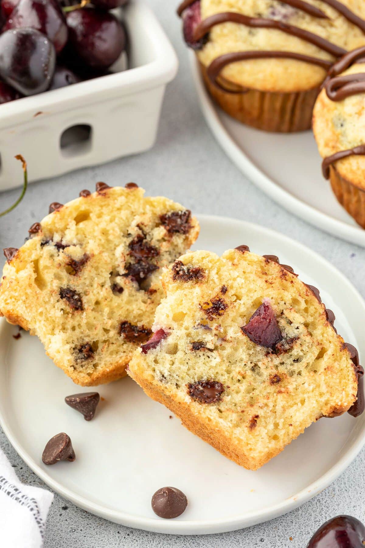 A muffin cut in half on a white plate, showcasing pieces of chocolate and cherries inside.