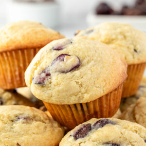 Several chocolate chip cherry muffins, stacked in a pile on a plate with cherries in a bowl in the background.