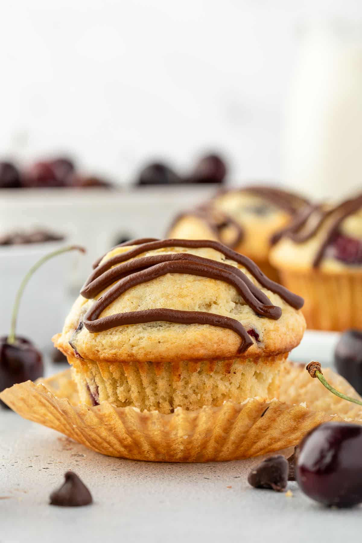 A cherry muffin with chocolate drizzle on top, partially unwrapped from its paper with cherries and more muffins in the blurred background.