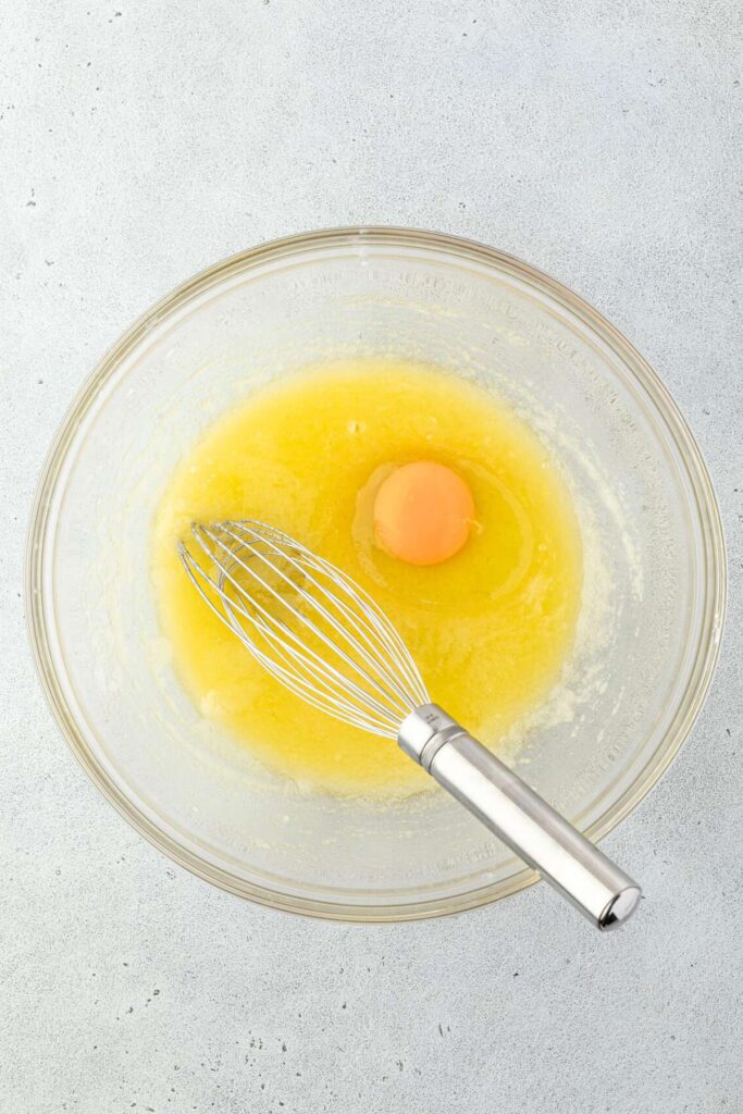 A glass bowl sits on a light surface, holding a whisk, melted butter, and a raw egg.