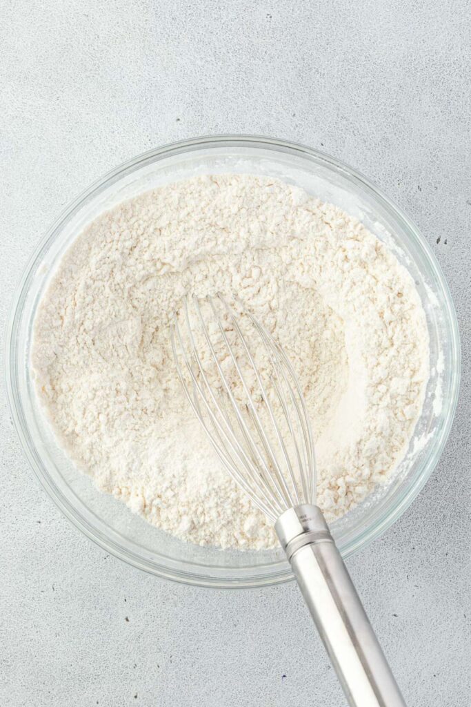 A glass bowl brimming with flour and a whisk sits on a light gray surface.