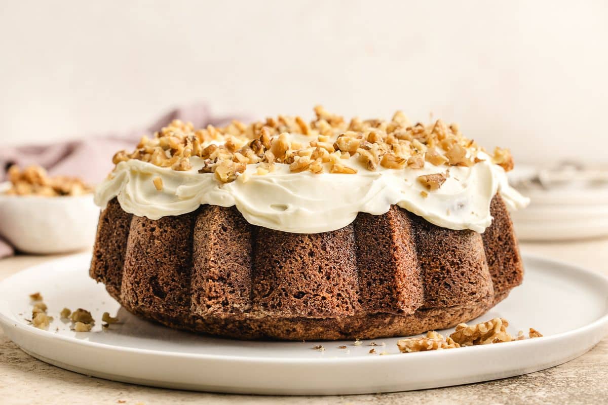 Carrot Bundt cake with cream cheese frosting and chopped walnuts on top, served on a white plate.