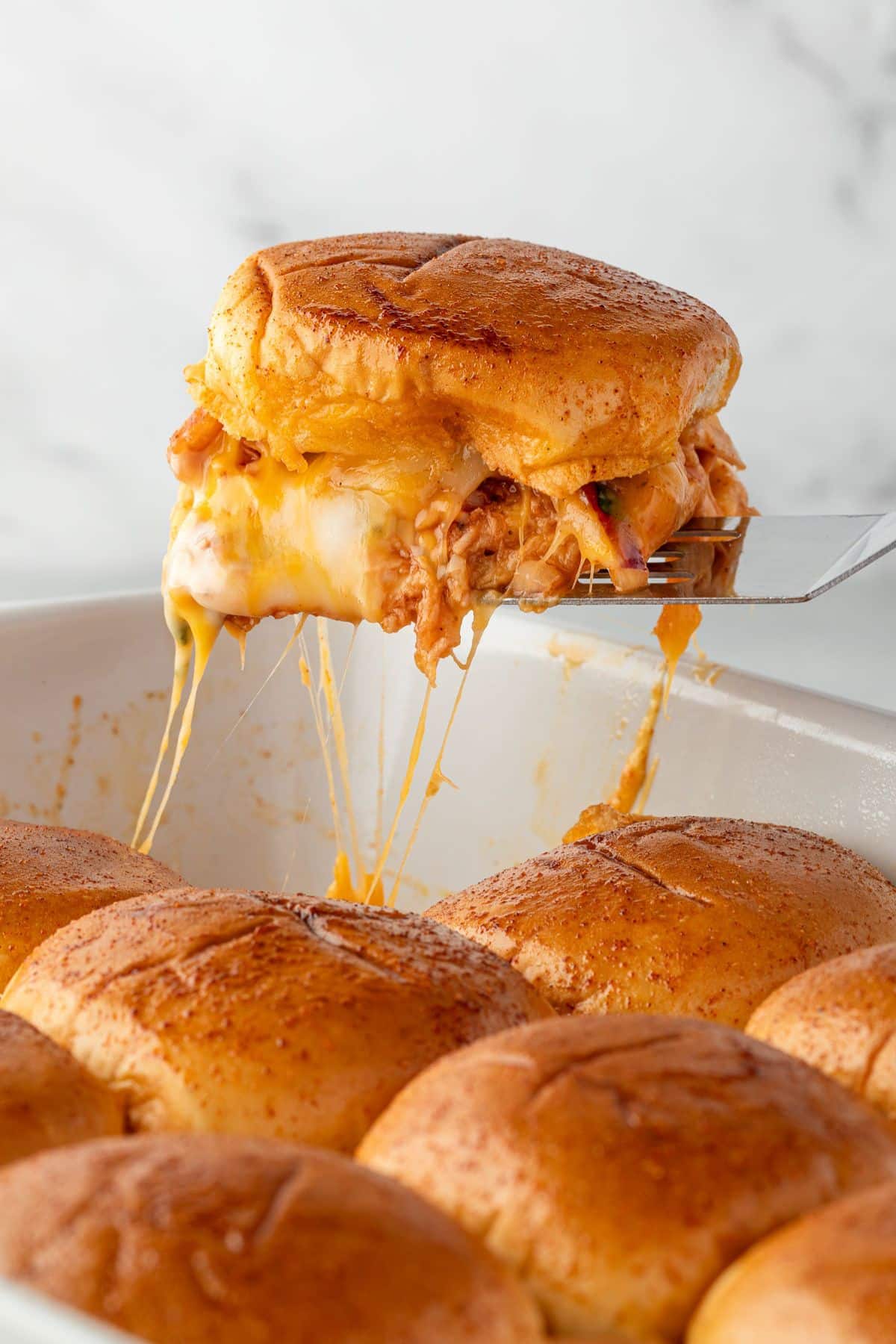 A cheesy pulled chicken slider being lifted from a baking dish of golden, baked sliders.