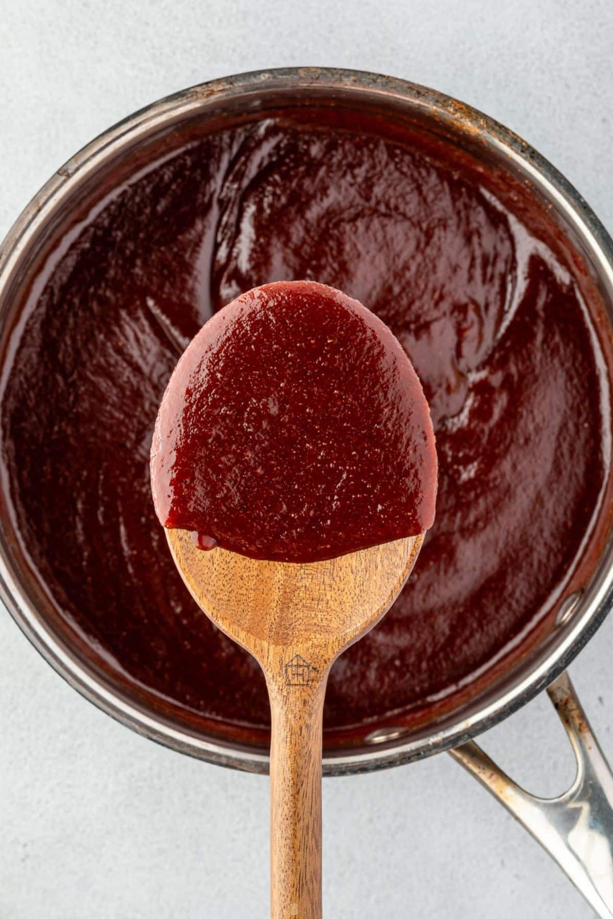 A wooden spoon holding thick barbecue sauce over a pot filled with more sauce.