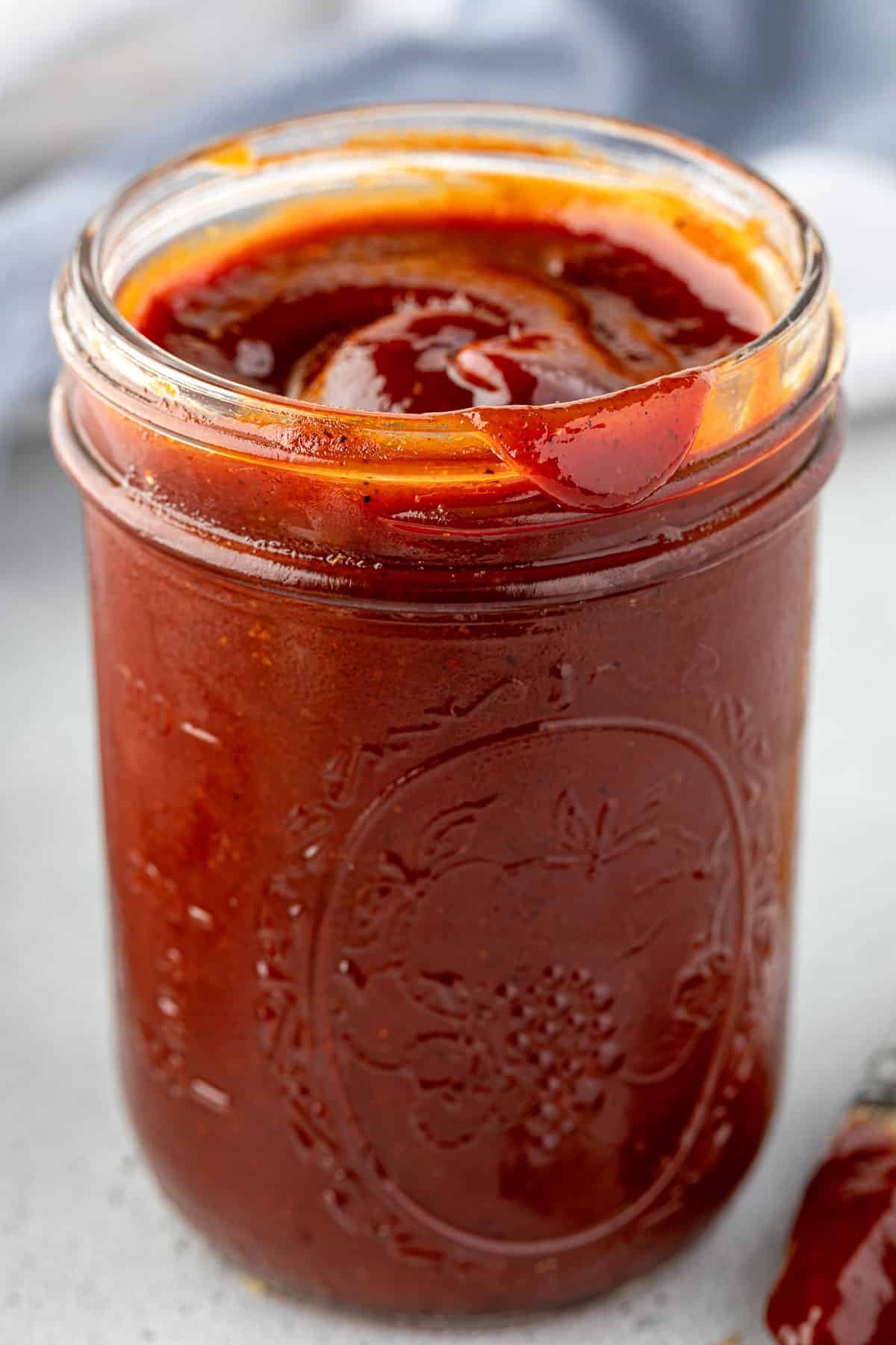 A glass jar filled with thick, red barbecue sauce on a light surface.