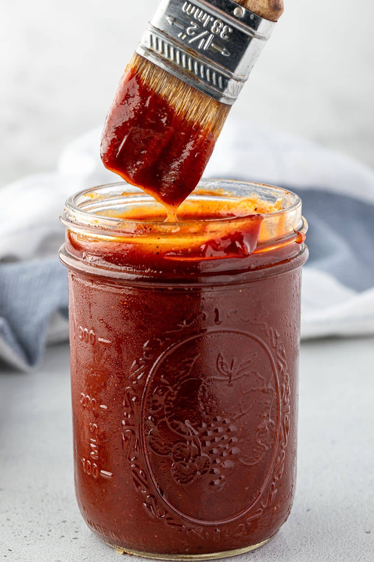A brush coated in barbecue sauce is held above a glass jar filled with more barbecue sauce.