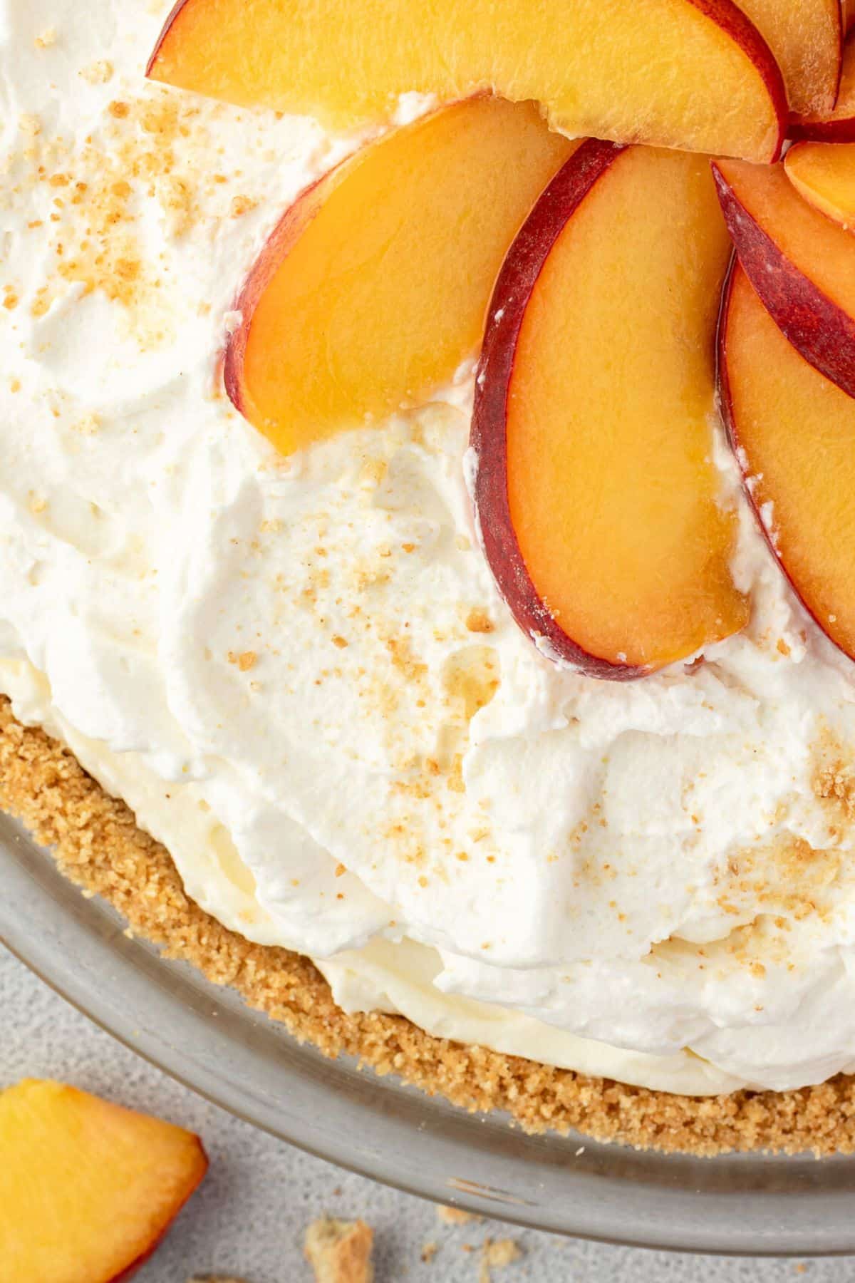Close-up of a pie with whipped cream, peach slices, and a crumbly crust in a glass dish.