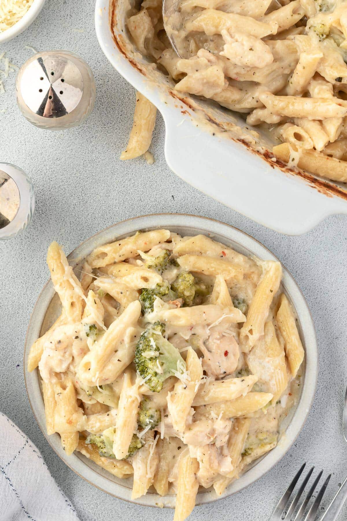 Plate of creamy baked penne pasta with chicken and broccoli, next to a casserole dish with more pasta.