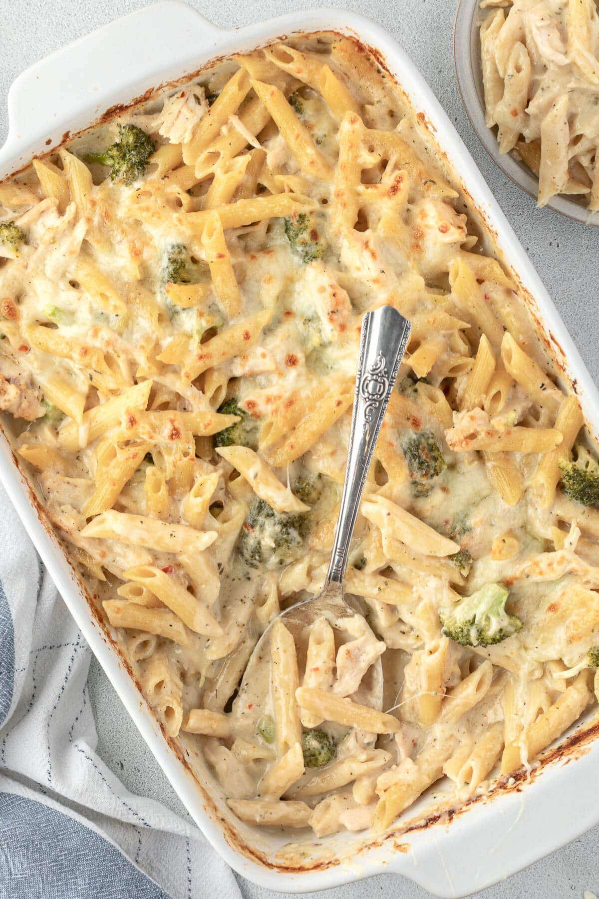 Baked penne pasta with broccoli, chicken, and cheese in a casserole dish, served with a metal spoon.
