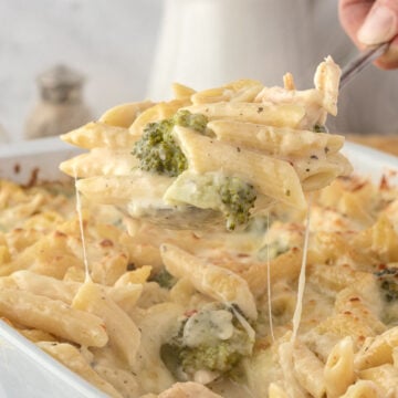 A spoon lifts cheesy baked penne pasta with broccoli from a casserole dish.