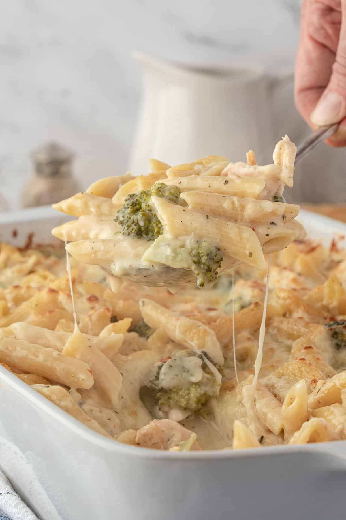 A hand lifts a cheesy pasta bake with broccoli from a white casserole dish.