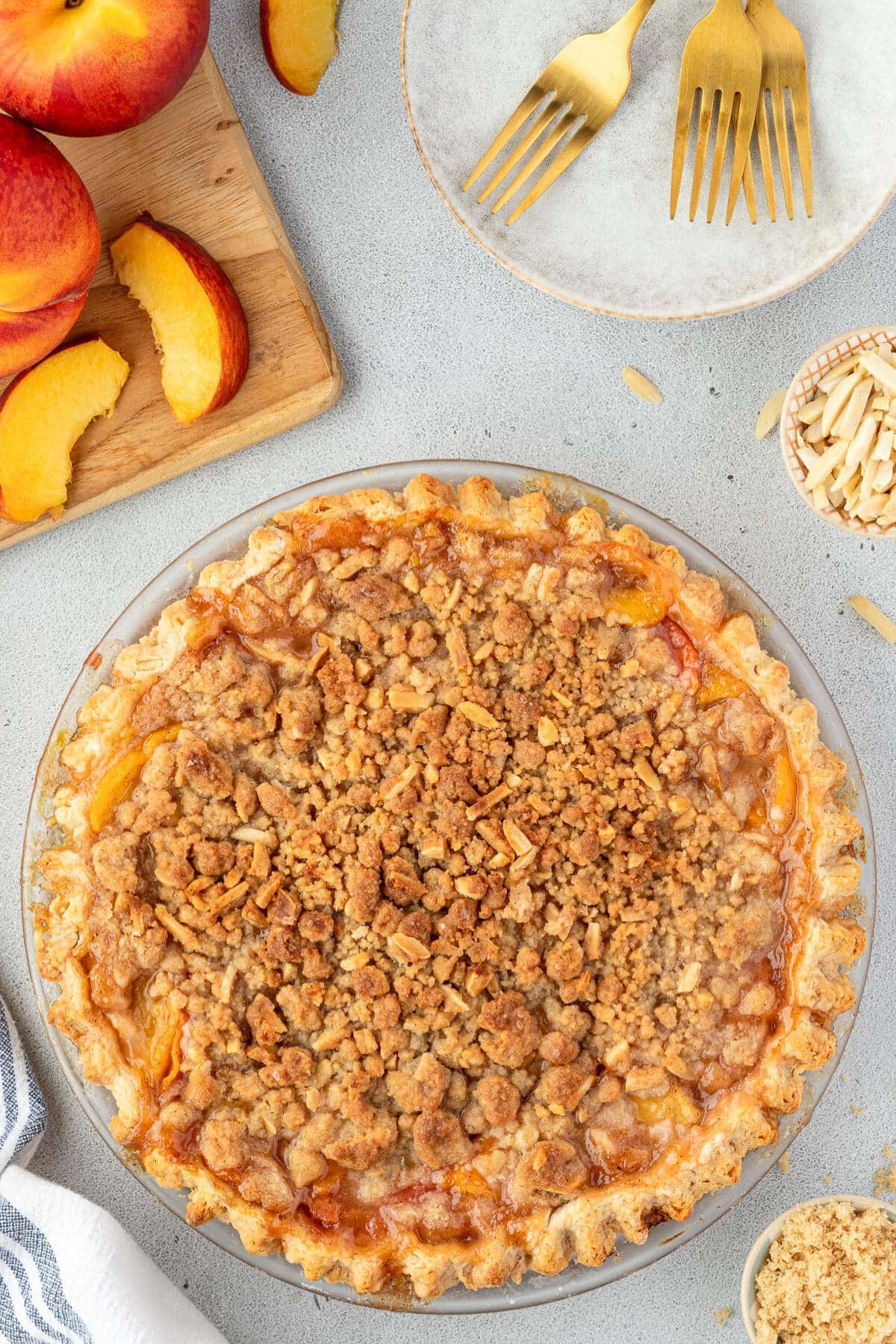 Peach crumb pie on a table with sliced peaches, almonds, two gold forks, and a plate nearby.